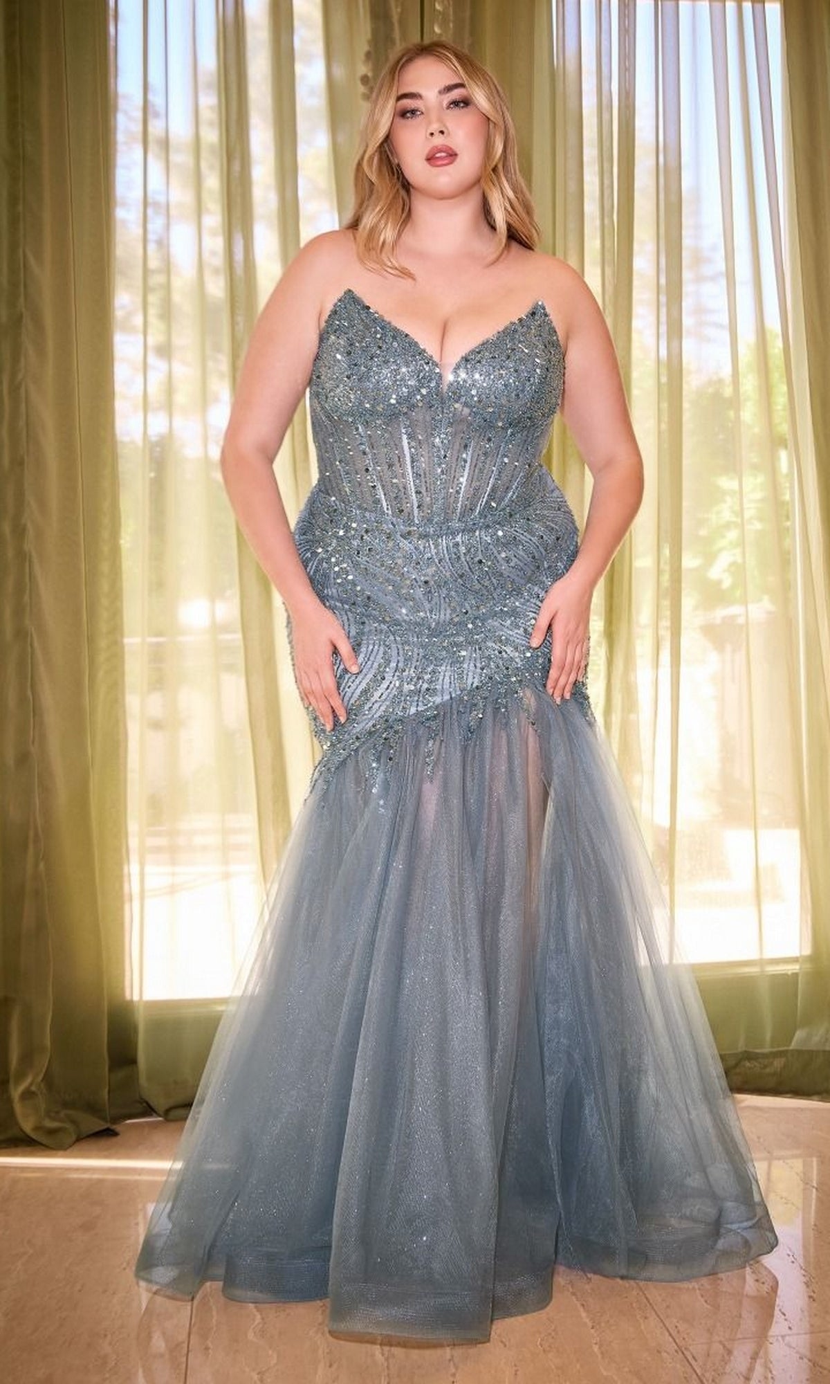 A woman stands indoors by sheer green curtains in sunlight, wearing the Plus-Size Long Mermaid Prom Dress CD0214C— a strapless, form-fitting blue sequin gown that shimmers beautifully.